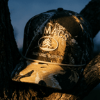 Camouflage cap with 'Macs Nature Supply' logo on a tree branch at night.