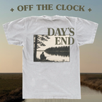 A white graphic T-shirt shown from the back, featuring a large printed scene of a person fishing by a lake with the words “DAY'S END.” The shirt is centered over a softly blurred outdoor landscape background, with “+ OFF THE CLOCK +” text above.
