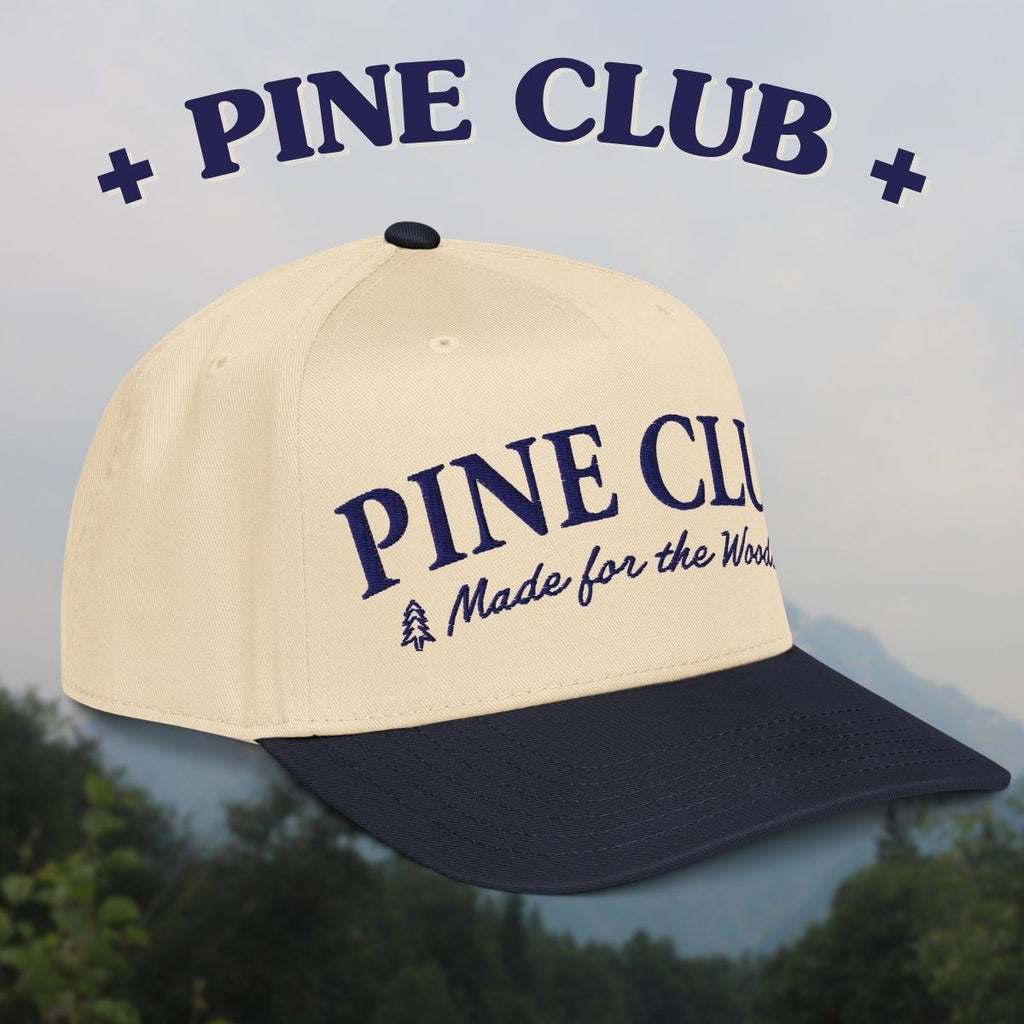 Beige and navy cap with 'Pine Club' branding against a blurred natural background