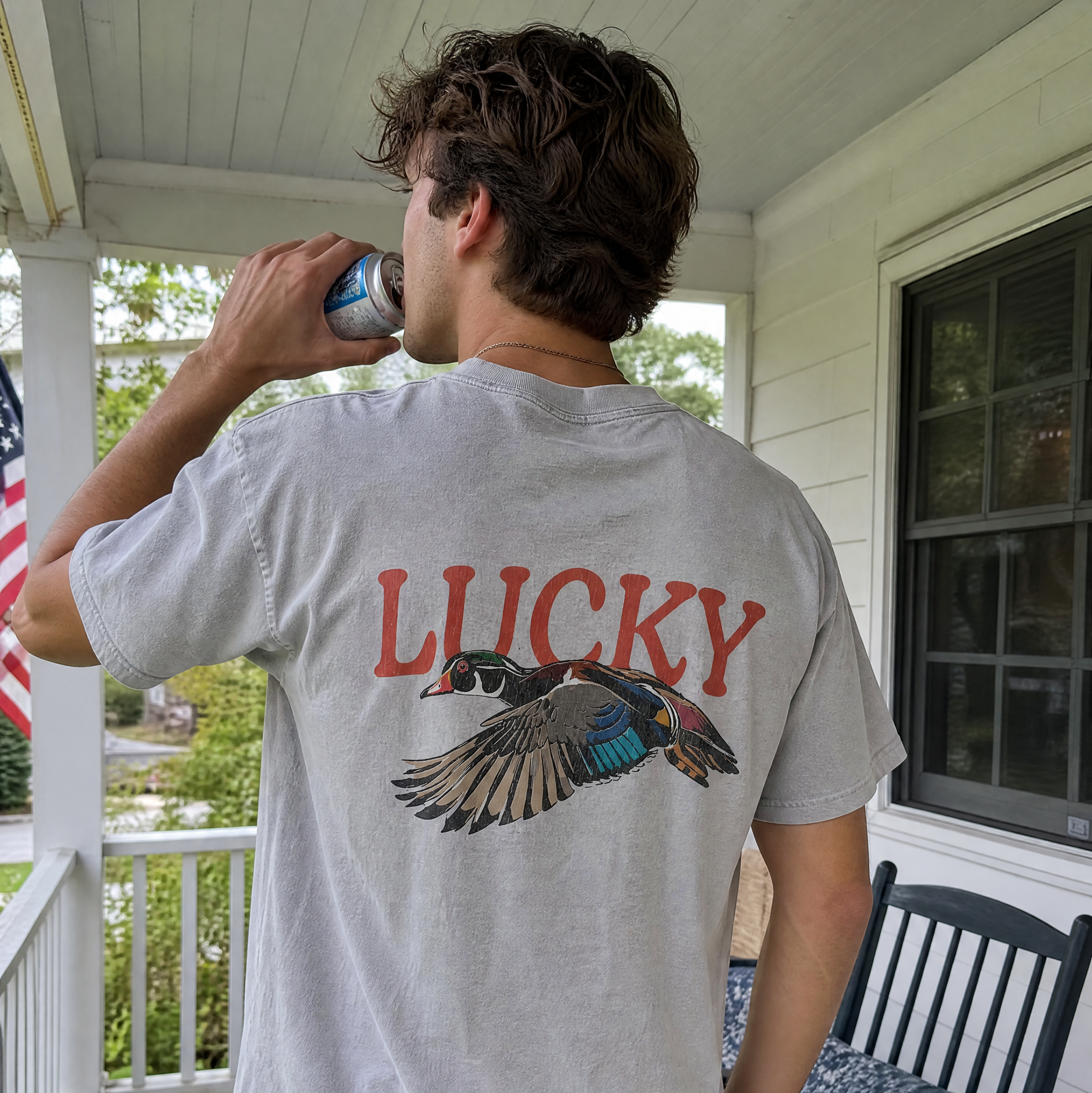 A man standing on a porch wearing a light gray t-shirt with a red 'LUCKY' text and a flying duck graphic on the back, paired with light blue jeans.